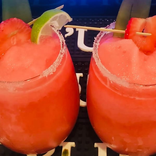 Two vibrant red frozen drinks, garnished with strawberries and lime, sit on a dark surface.